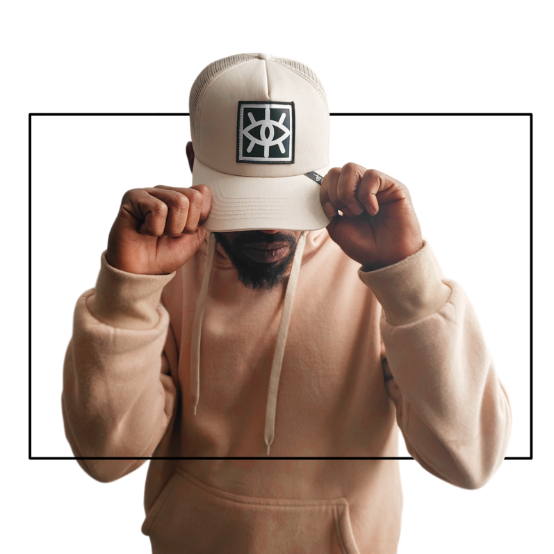 Hombre con gorra clara y sudadera, inclinado hacia adelante, posando con las manos en la gorra.